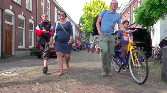A family walks four abreast down a brick-paved street. A man wearing a blue shirt in the center walks a yellow bicycle