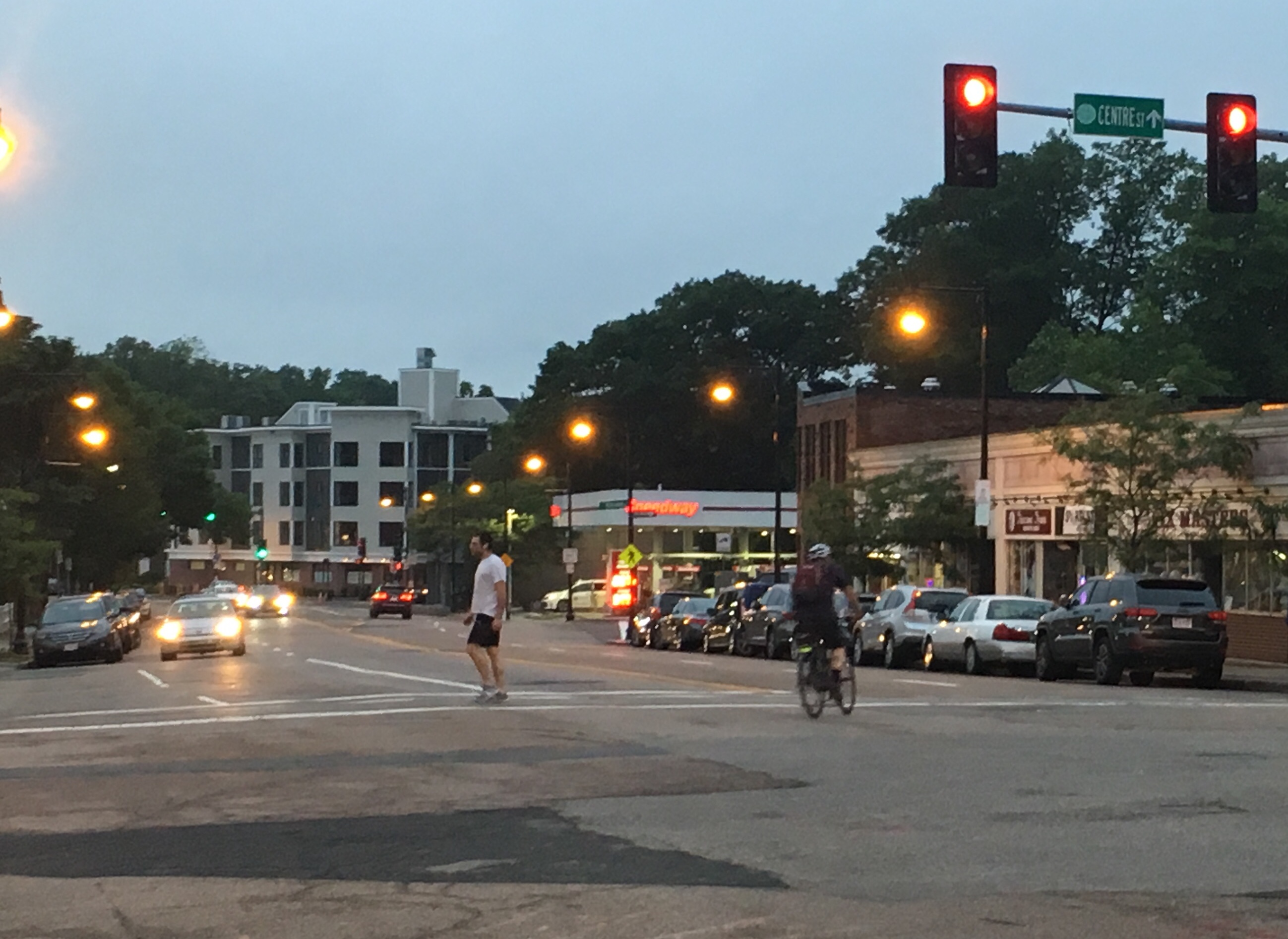 An evening scene on a wide four-lane street that's lined with one-story commercial buildings. In the foreground, a person walks across the street at a traffic light as another person rides a bike in the opposite direction. A few cars' headlights are visible in the background.