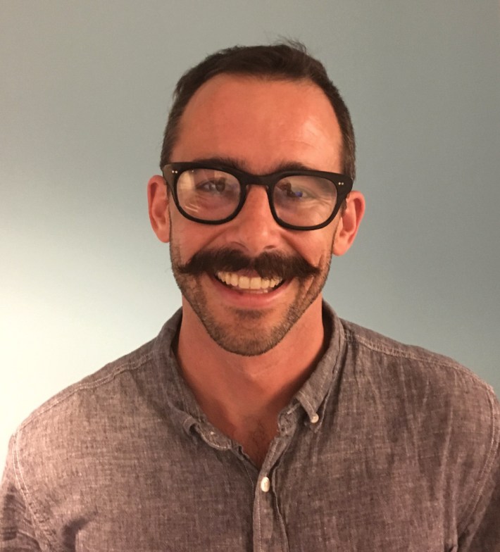 A headshot of Christian MilNeil, who is wearing glasses and a collared blue shirt