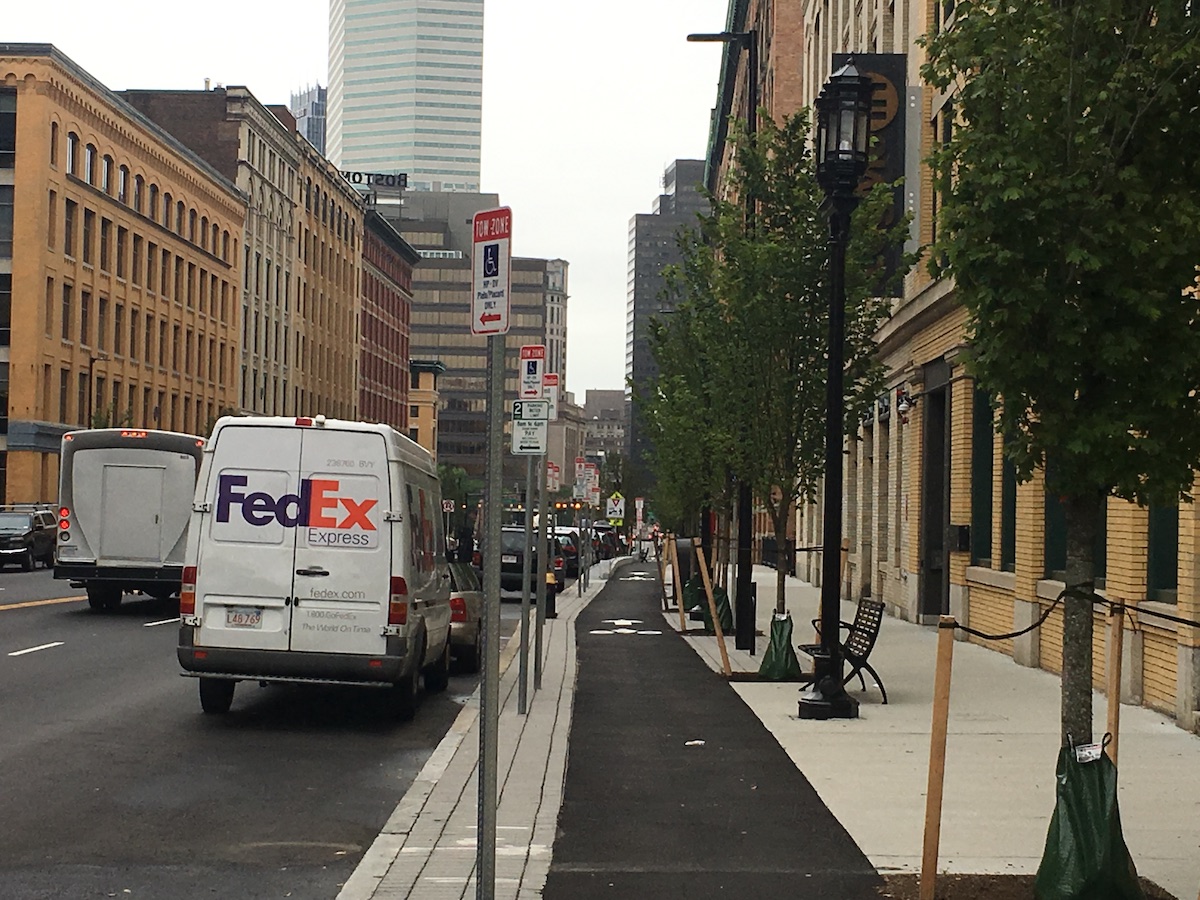 A view of a sidewalk-level bike lane running alongside newly planted sidewalk trees through a neighborhood of historic brick buildings. In the distance a downtown Boston skyscraper is visible.