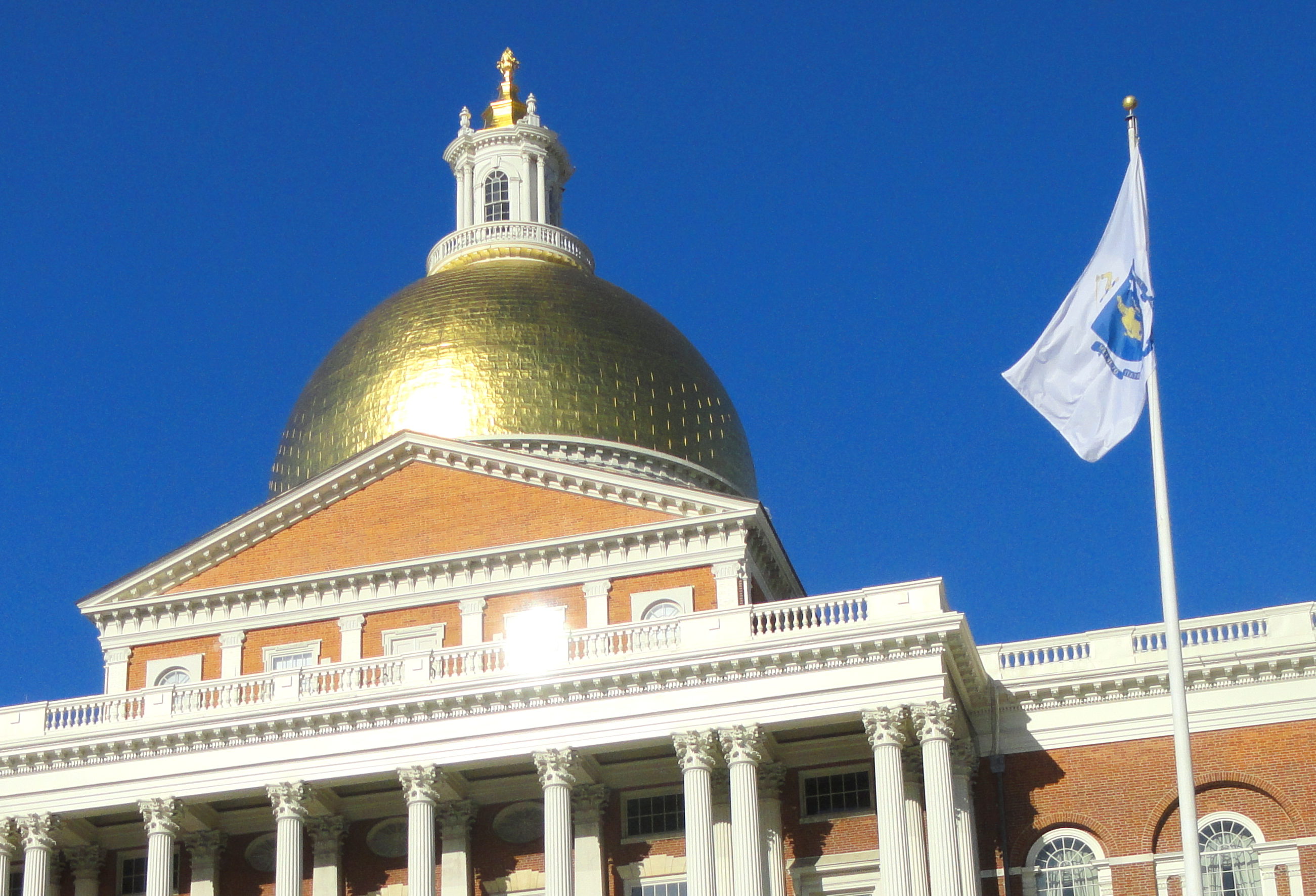 The gold-plated dome of the Massachusetts State House against a blue sky, with the Massachusetts flag flying to the right of the dome in the foreground.