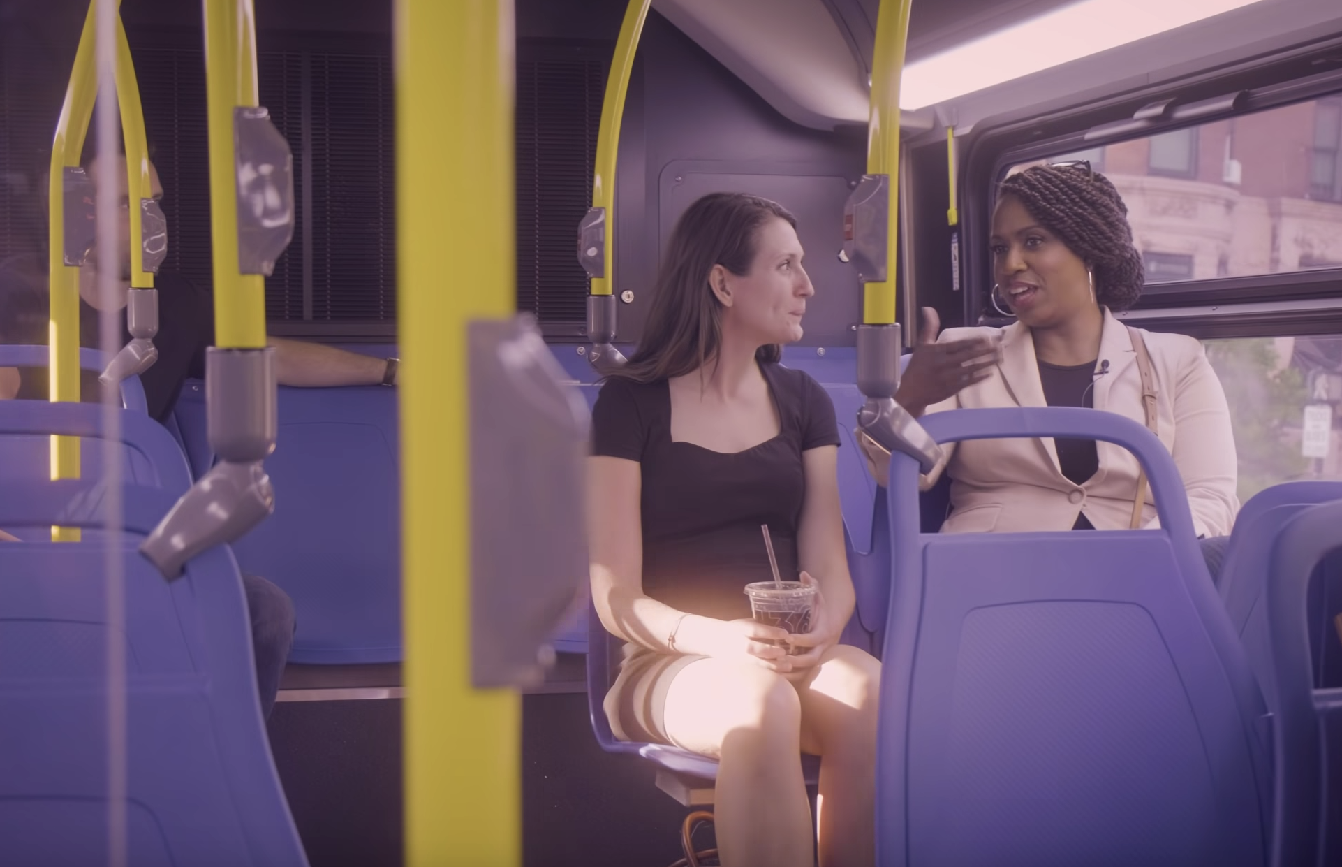 A Black woman gestures while talking with a white woman seated next to her on a city bus.