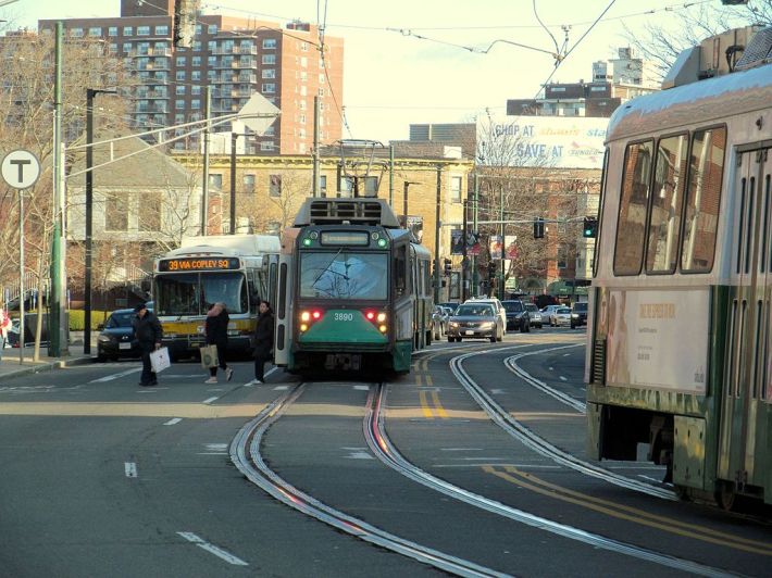 Two Green Line trains meet at the Mission Park stop of the E Branch on Huntington Ave. in Boston. This segment of the Green Line is one of the last remaining areas where trains must share a lane with car and truck traffic, and stations lack dedicated platforms where riders can board and disembark. Photo by Wikimedia Commons user Pi.1415926535, used with permission under a Creative Commons license.