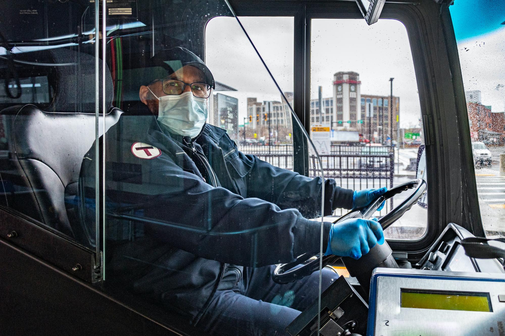A bus driver wearing a face mask and a jacket with an MBTA "T" logo on his arm sits behind the wheel of a bus.