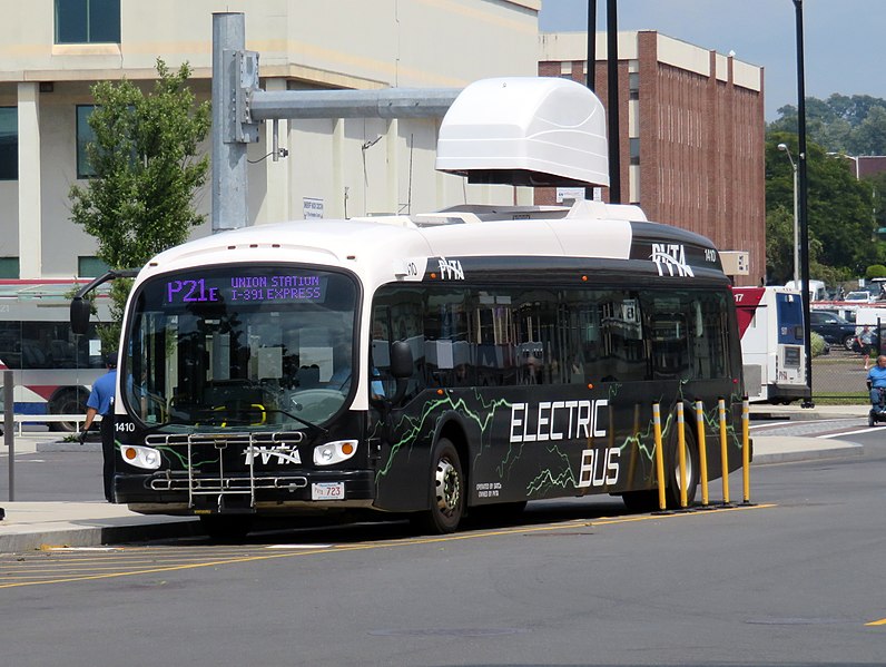 A black bus with the words "ELECTRIC BUS" on its side parks in a parking lot under a white charger, which is supported by a large steel mast arm that's cantilevered over the bus. In the background are some city buildings.