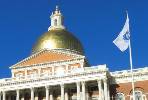 Front view of the Massachusetts State House with sunlight glinting off the gold dome at the top and a Massachusetts state flag at right.
