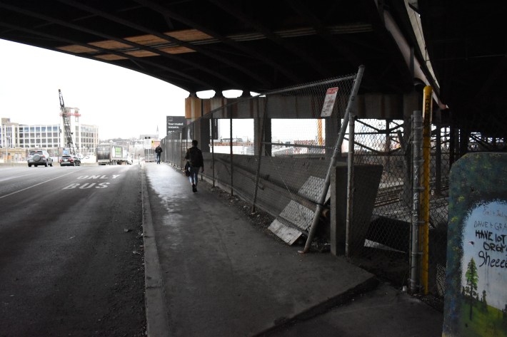 Pedestrians walk along the narrow sidewalk on Maffa Way, an off-ramp from I-93 and the primary walking route between Sullivan Square Orange Line stop and East Somerville.