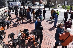 A crowd gathers in front of the Massachusetts State House next to a handful of pedal-assist electric bikes.