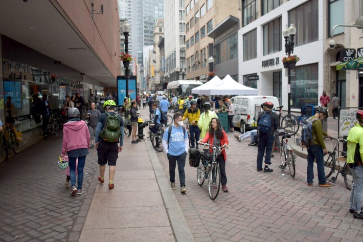 A crowd of Bike to Work Day celebrants, many of whom are wearing bike helmets, gather in front of tents to get free coffee and pastries in Downtown Crossing.