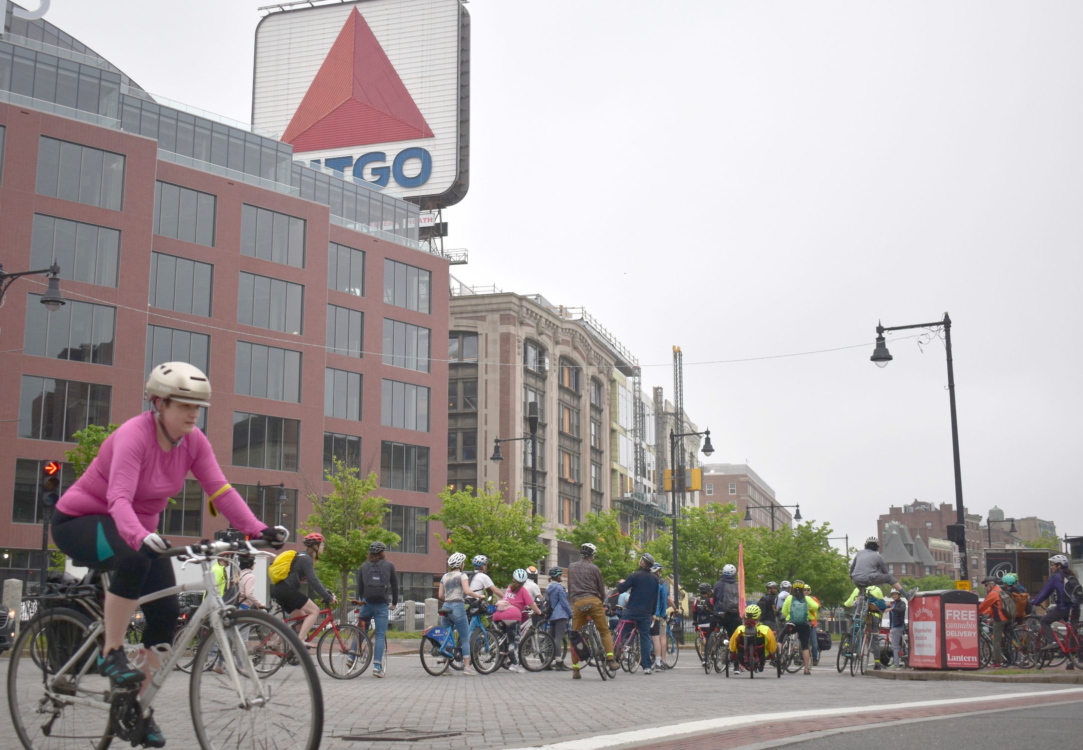 A large crowd of bike riders gathers on Commonwealth Avenue in Kenmore Square beneath the Citgo sign.