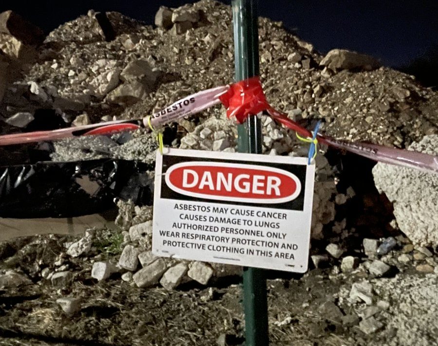 A hazardous materials warning sign reading "DANGER: Asbestos may cause cancer, causes damage to lungs, authorized personnel only, wear respiratory protection and protective clothing in this area" hangs from a post in front of an uncovered pile of construction debris.