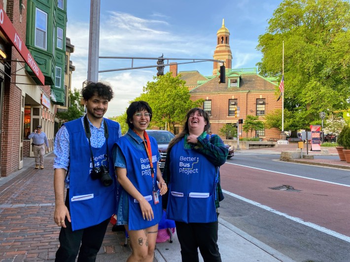 Three "bus network redesign" outreach staff wearing blue aprons the the MBTA's "Better Bus Project" logo and pockets full of handout flyers smile on the sidewalk in front of Chelsea City Hall next to a red bus lane.