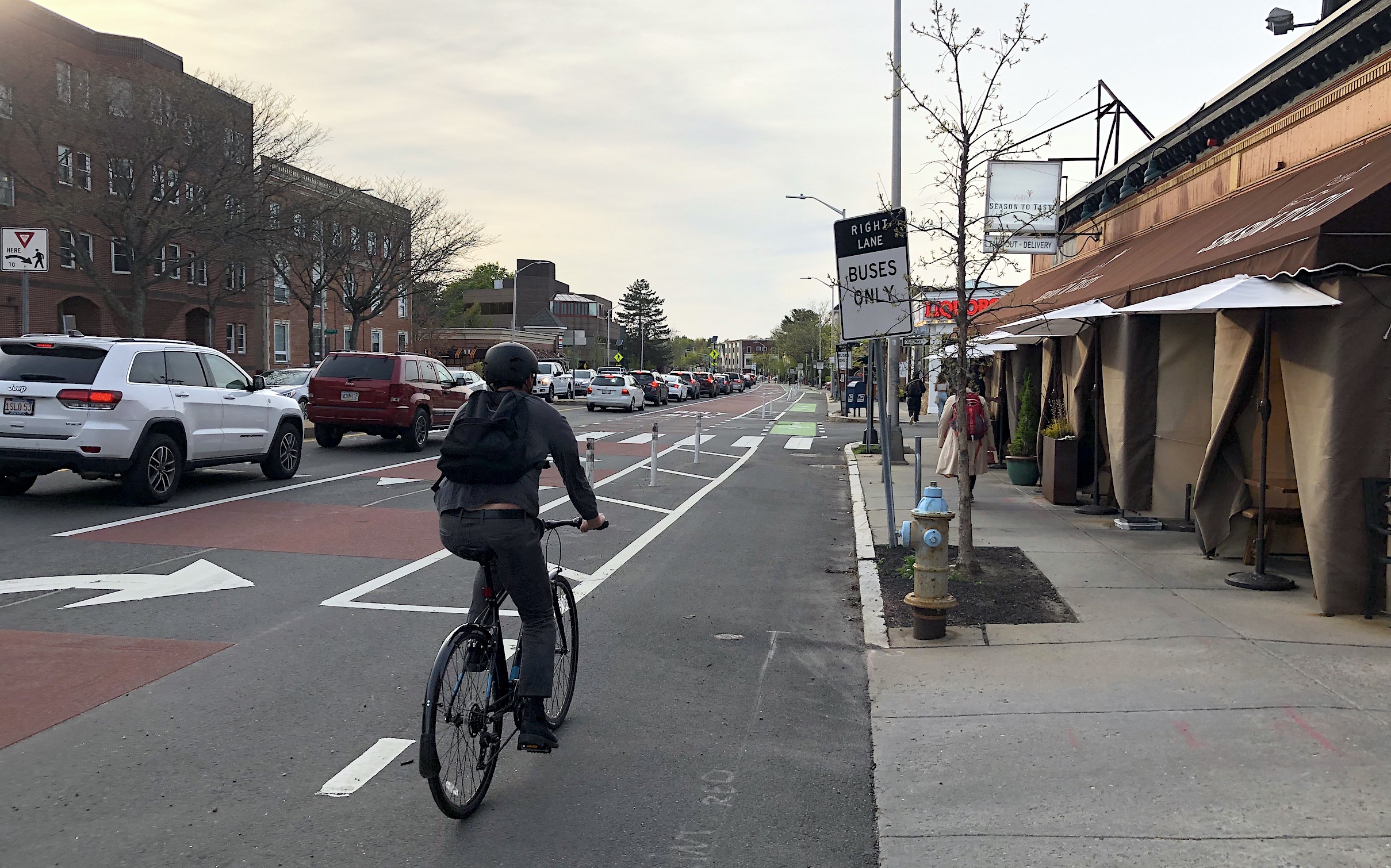 A bike rider rides along a flexpost-protected bike lane next to a sidewalk cafe on one side and a line of cars waiting in traffic on the other side of the bus lane, on the left edge of the photo.