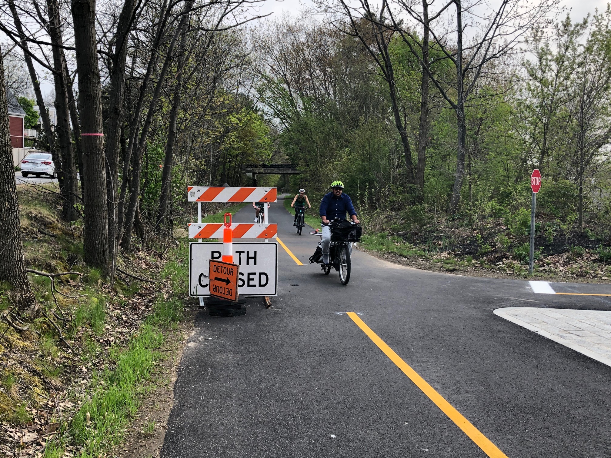 A "Path closed" sign is obscured by a construction cone and "detour" sign at the northern end of the Watertown-Cambridge Greenway in Cambridge while three people on bikes ride past.