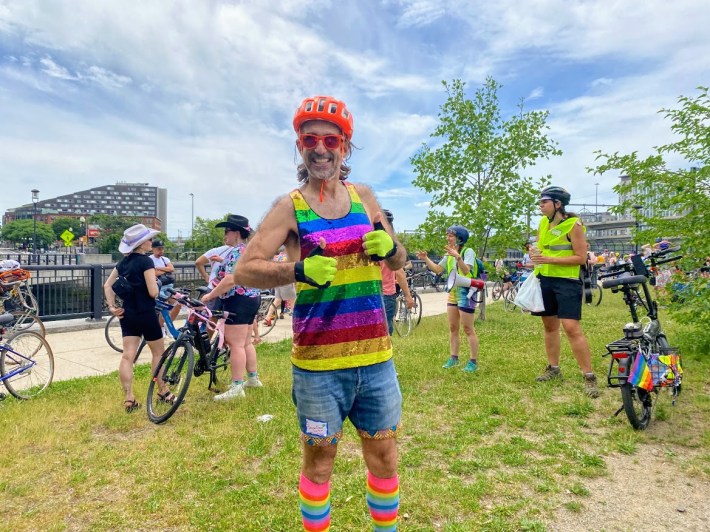 DJ Pussy Willow wearing a colorful pride tank top and socks, posing with a crowd of bicyclists behind him