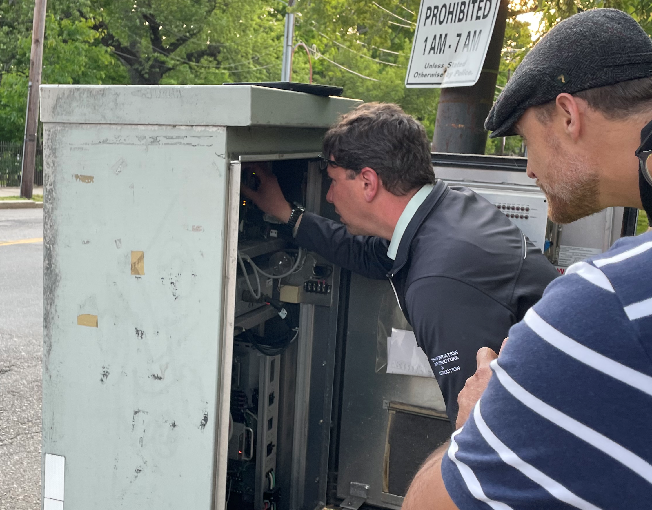 Dept. of Conservation and Recreation Chief Engineer Jeff Parenti leans into a metal traffic signal cabinet on Broadway in Arlington to adjust the traffic signal timing of the pedestrian crosswalk while Somerville Director Transportation & Infrastructure Brad Rawson watches over his shoulder.