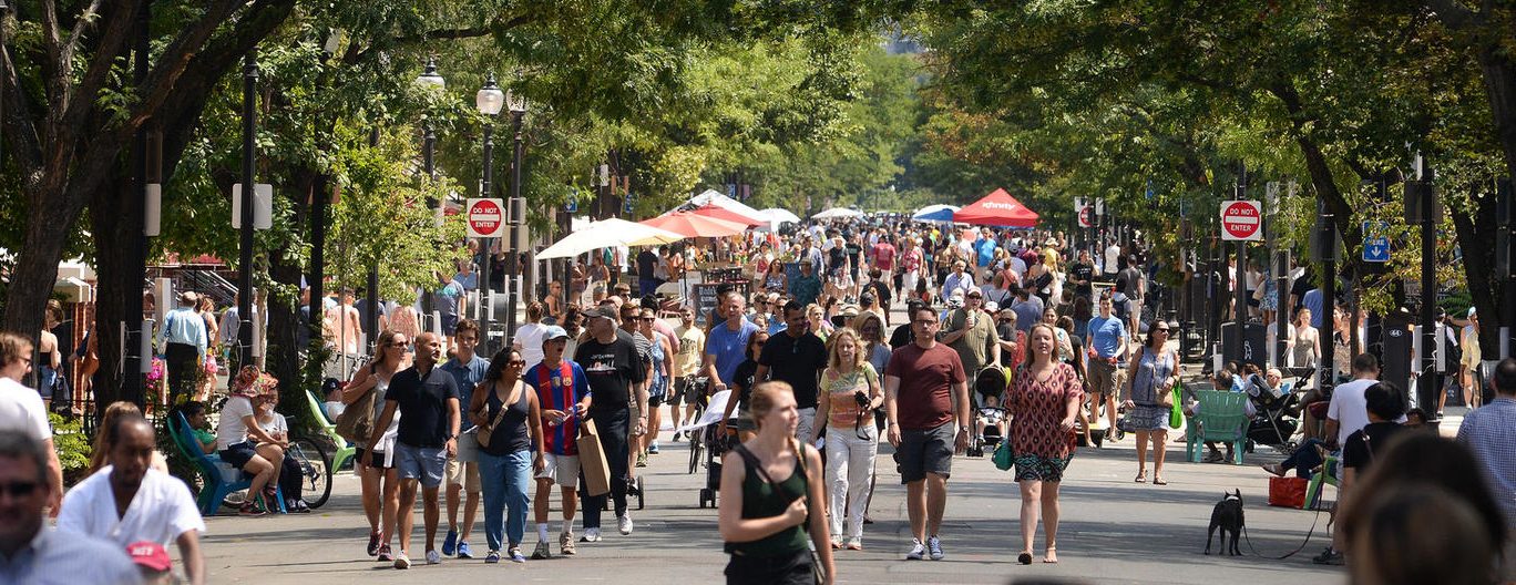 Crowds walk down the middle of a car-free Newbury Street among vendors' tents, umbrellas for outdoor dining, and people lounging in lawn chairs during one of the city's popular Open Streets events.