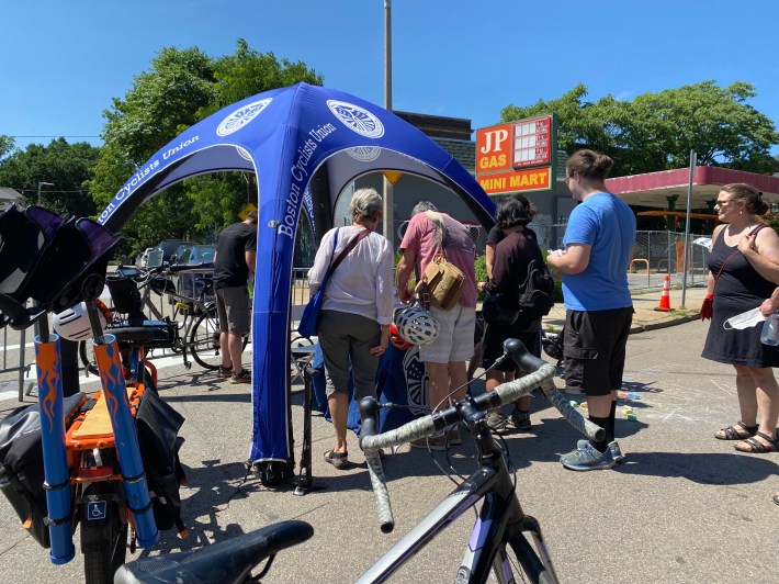 A crowd gathers around the Boston Cyclists Union bike repair tent in front of an abandoned gas station.
