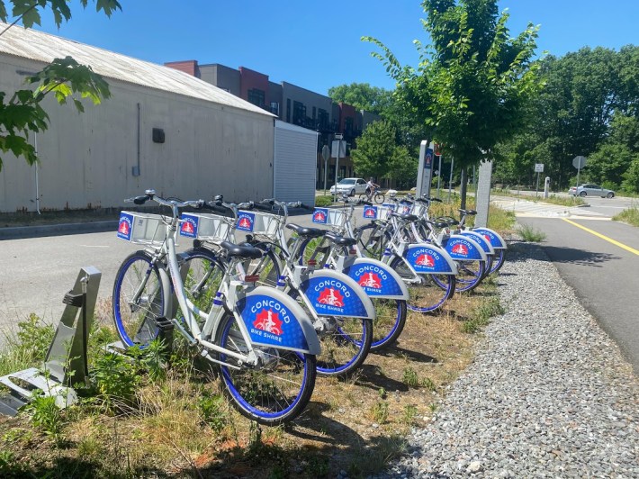 A row of six gray-and-blue bikeshare bikes parked in the grass next to a bike trail.