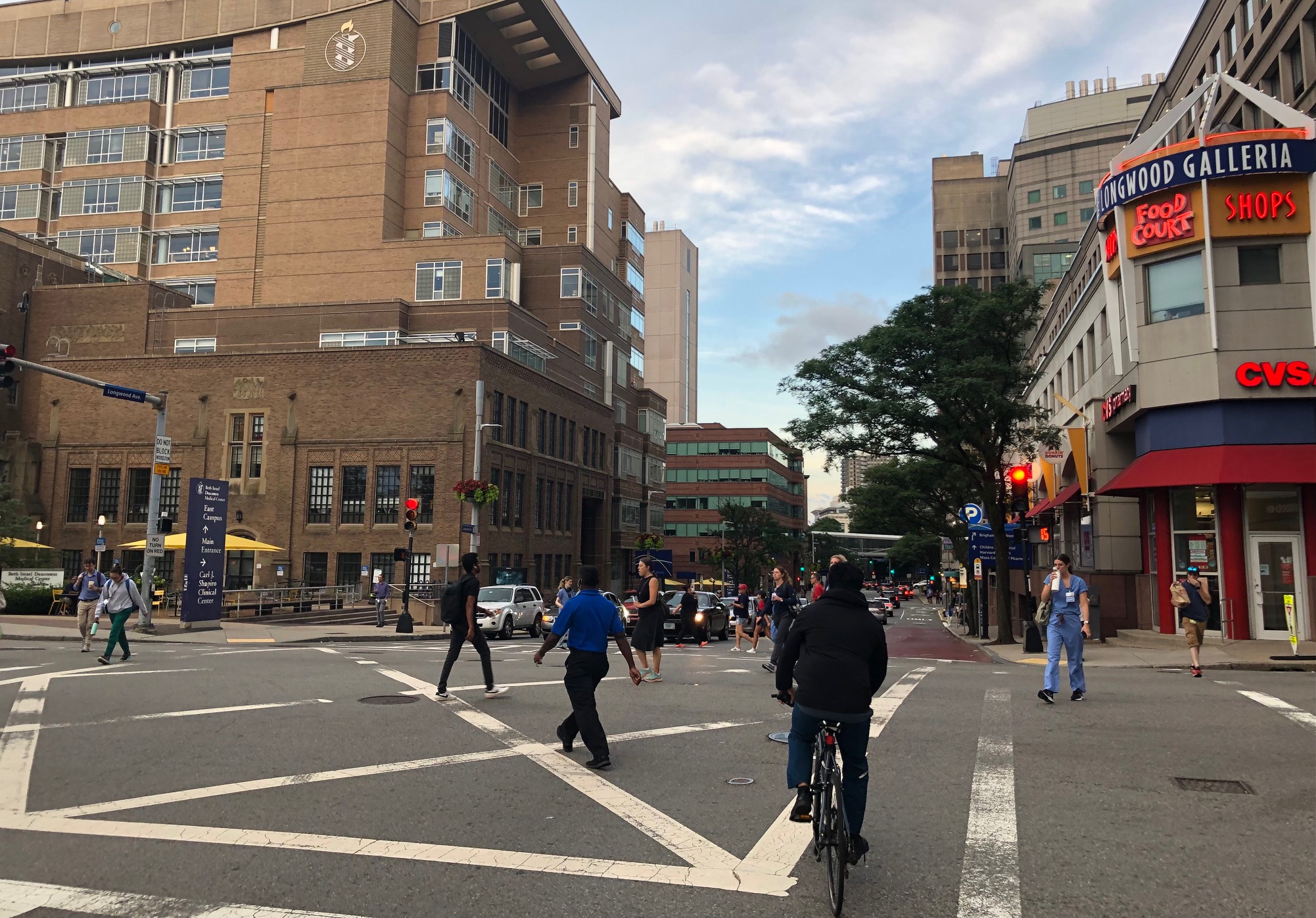 A dozen people walking in all directions, plus one person on a bicycle, crossing the intersection of Brookline and Longwood Avenues under the Longwood Galleria neon sign. The Longwood Area's skyline of hospital buildings is visible in the background.