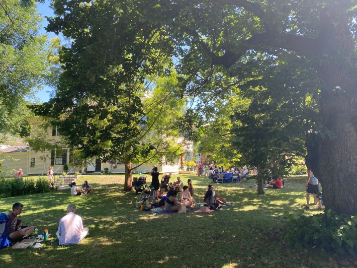 Groups of people relaxing on a shady lawn under big trees in front of a historic house