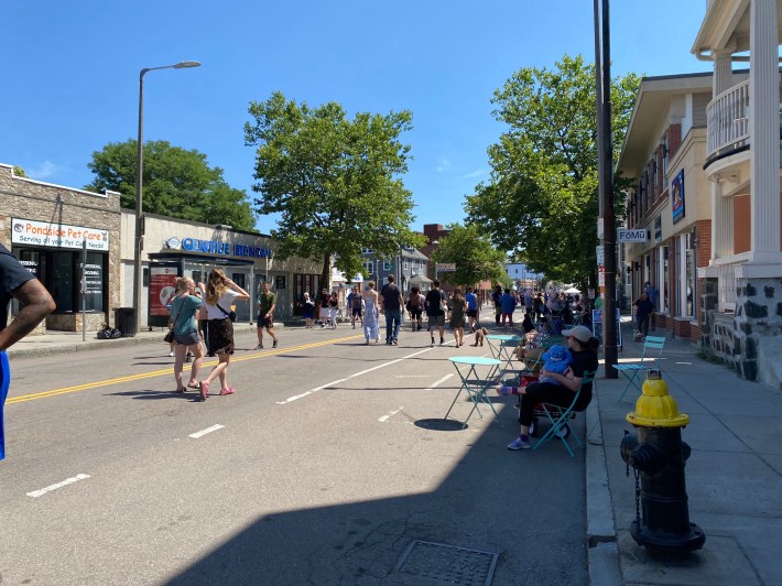 People relax at tables and chairs in the shade line the street where cars would typically park while people on foot and on bikes go by in the middle of the street.