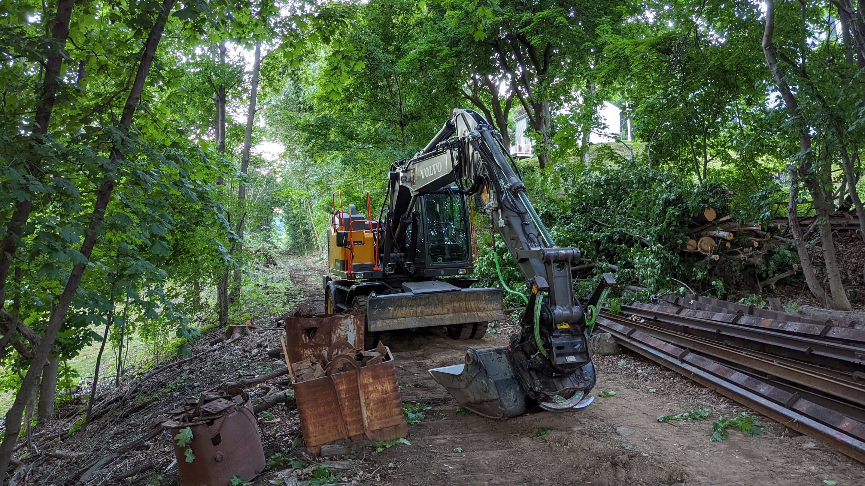 A backhoe rests on top of old railroad ties next to a pile of old rails along an abanoned railroad corridor surrounded by leafy trees.