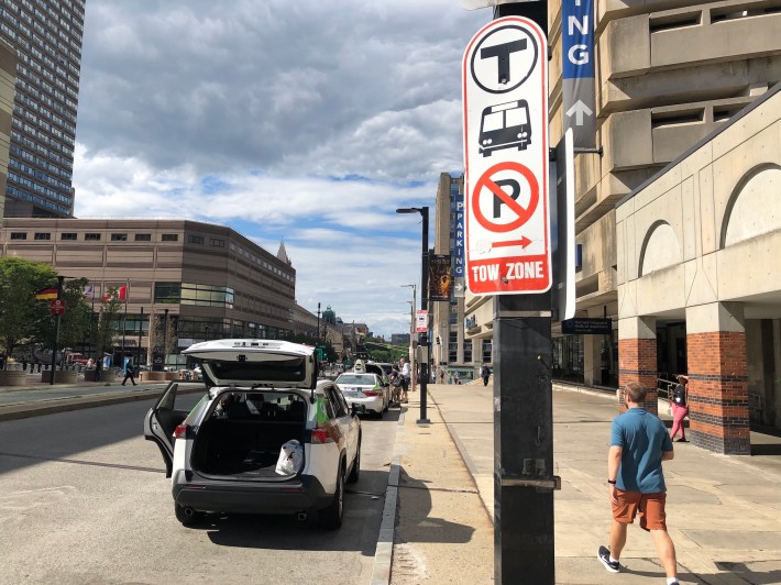 A "no parking tow zone" sign sits along side a new shuttle bus loading zone outside Back Bay Station on Dartmouth Street in Boston. A dozen taxis are parked in the loading zone.