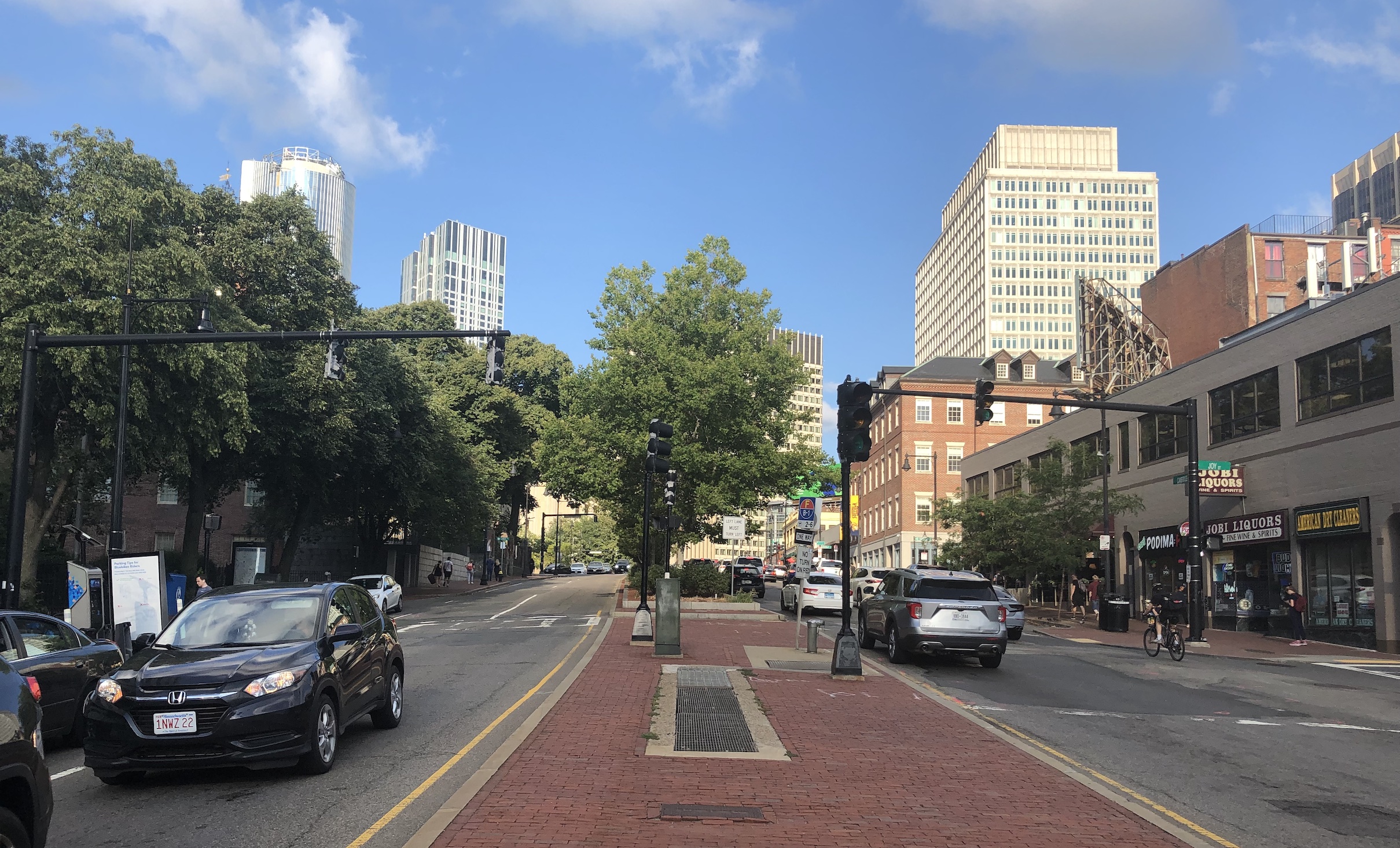 Looking east up Cambridge Street towards Government Center near the intersection of Joy Street. A wide brick-paved median dominates the foreground view; to the sides are two lanes of car traffic in each direction. A bike rider climbs the hill on the right edge of the photo.