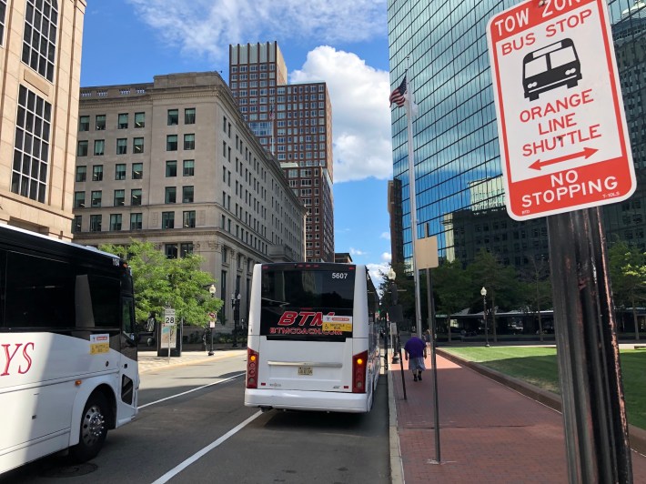 Two shuttle buses on training runs pass a "tow zone - Orange Line shuttle bus stop" sign on Clarendon Street next to Trinity Church in Copley Square.