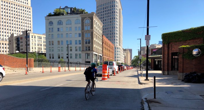 A bicycle rider enters a bike lane delineated with cones and construction barrels on Columbus Avenue in Back Bay. The bike lane runs along the curb, with a row of parked cars to its left.