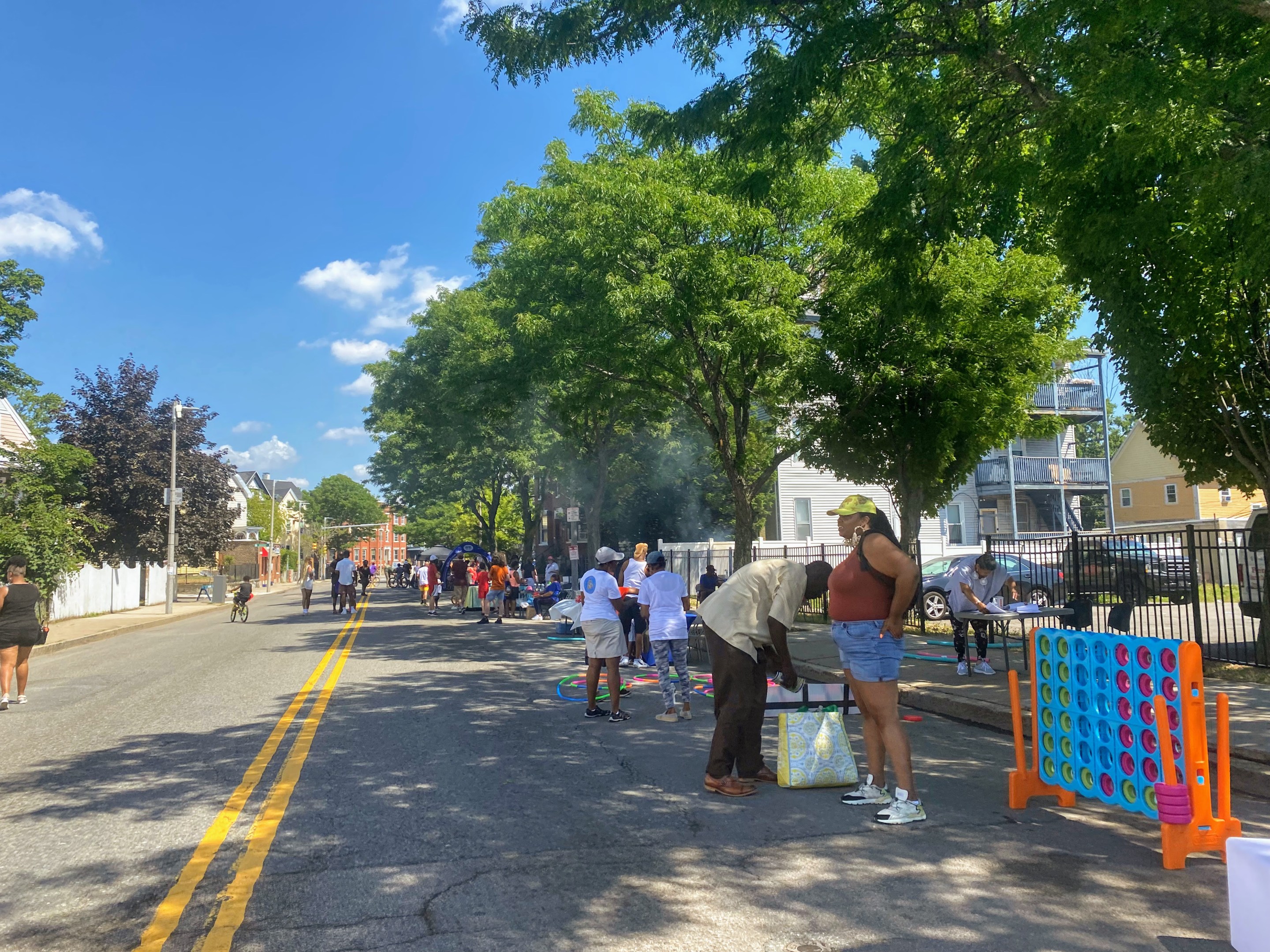 Groups of people chat with each other, ride bikes, and play games in the shade of several leafy trees on a street that's been closed to cars.