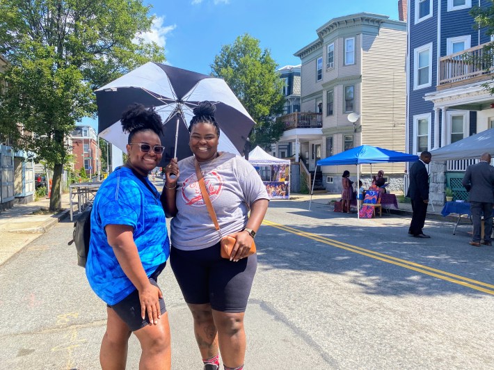Chelsea Jones (left) and Janessa (right) pose together under the shade of an umbrella during a hot Saturday morning at Open Streets Roxbury on Blue Hill Avenue. Janessa shared she likes this kind of programming coming to communities. “We need programming in this area, so I definitely think that it's good because it's kind of like forcing community in a way. It forces the people that are here to create community, come out, go to a vendor, meet people..” she said.