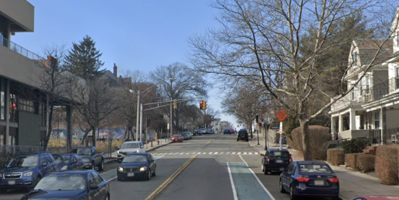A Google Street View image of Broadway in Somerville near the site where Stephen Conley crashed. The roadway includes two parking lanes against the curbs, two travel lanes for cars, and two green-painted bike lanes in between the parking and moving vehicle lanes. In the middle distance is a traffic light at the intersection of Packard Avenue. The bike lanes do not continue beyond that intersection.