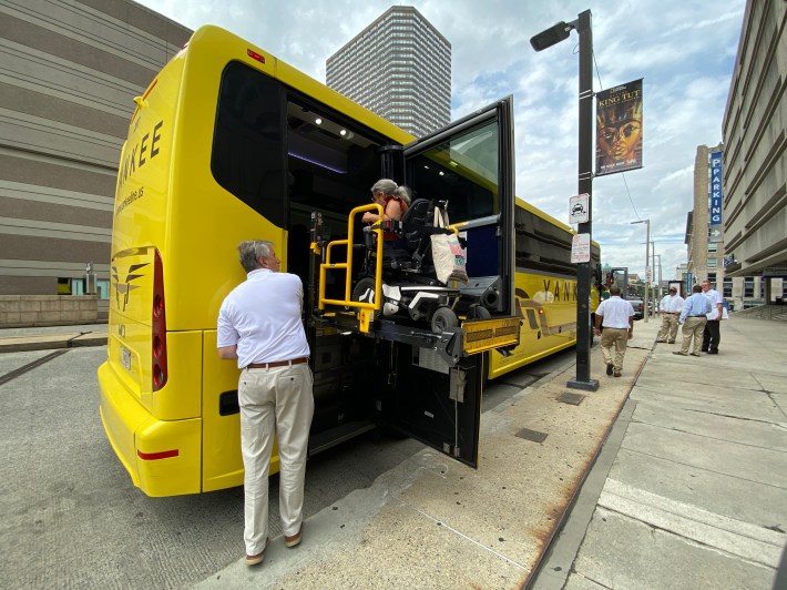 A wheelchair user rides on a lift to board a yellow Yankee Line bus from its rear door while an attendant assists from the sidewalk.