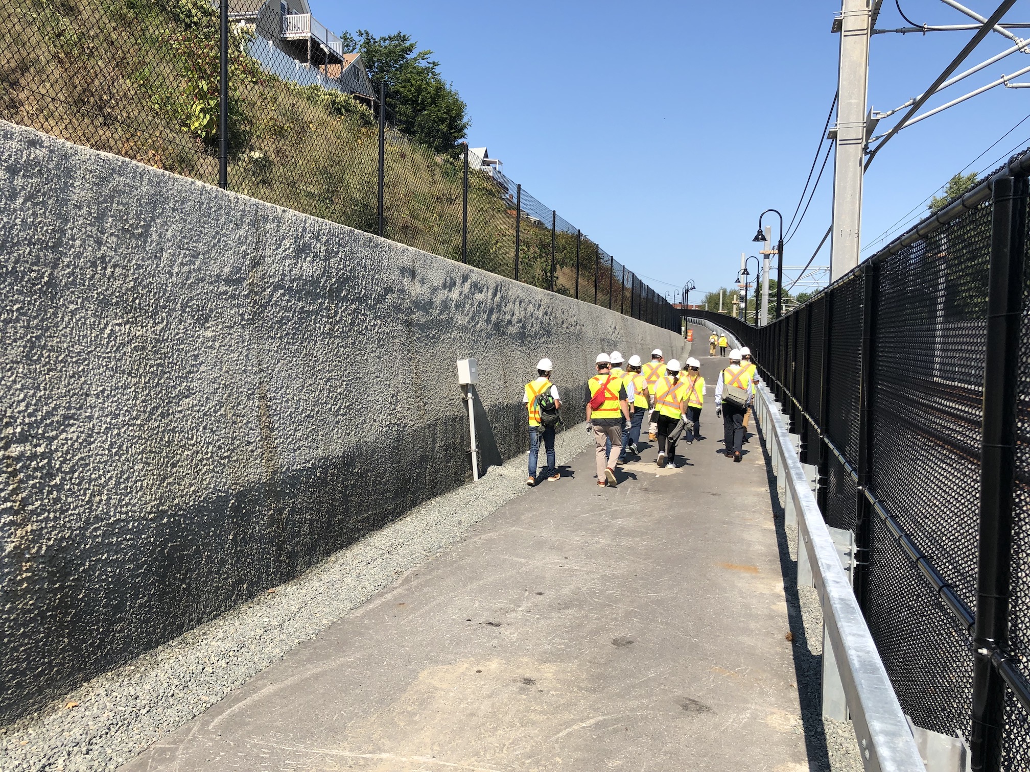A crowd of people in construction vests climbs a paved path as it gently climbs uphill next to a retaining wall (left) and a fence that separates the trail from the Green Line tracks (right)