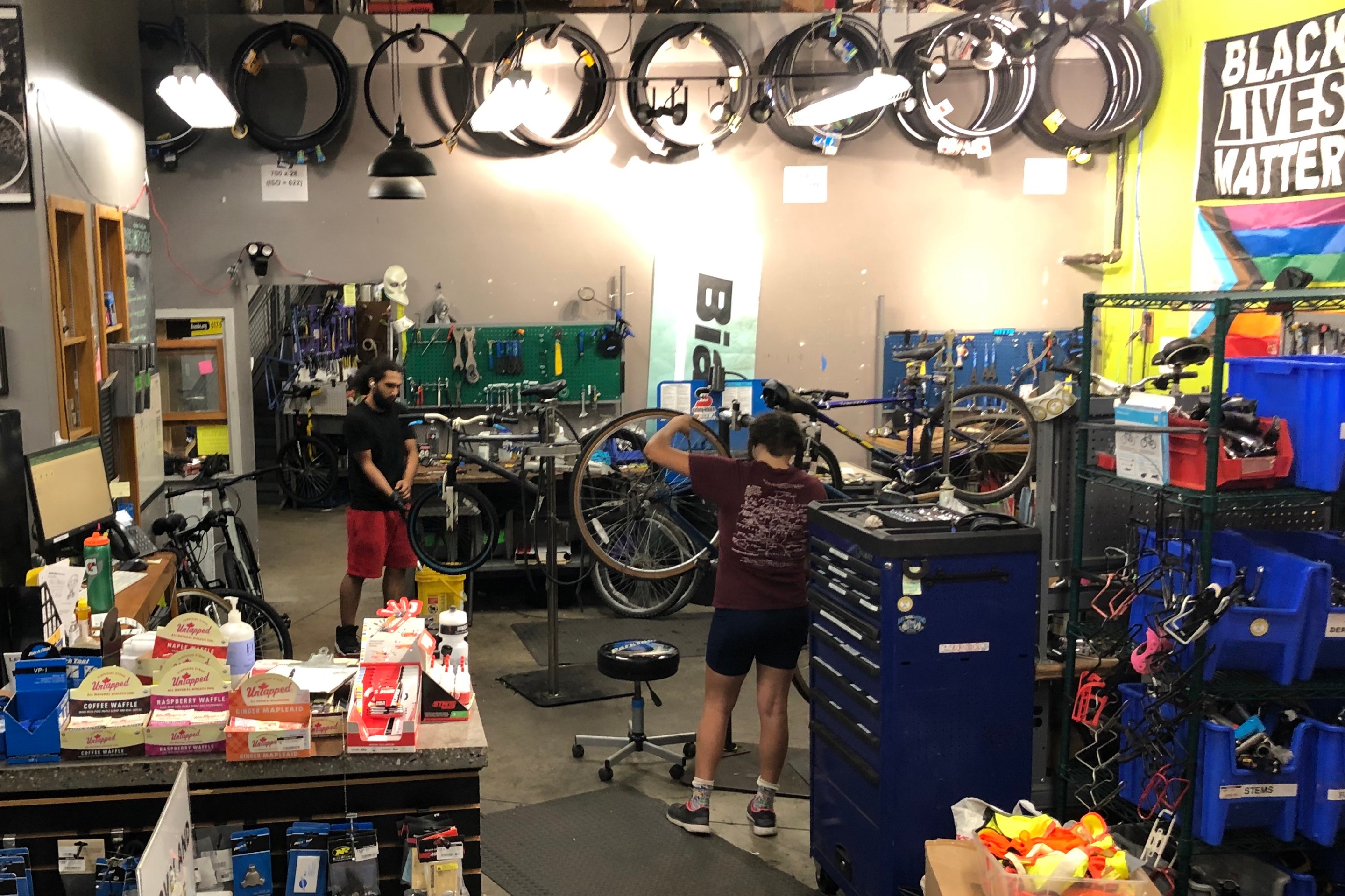 Two mechanics workon bikes on stands in a crowded workshop with bike tires hanging from the high ceilings.