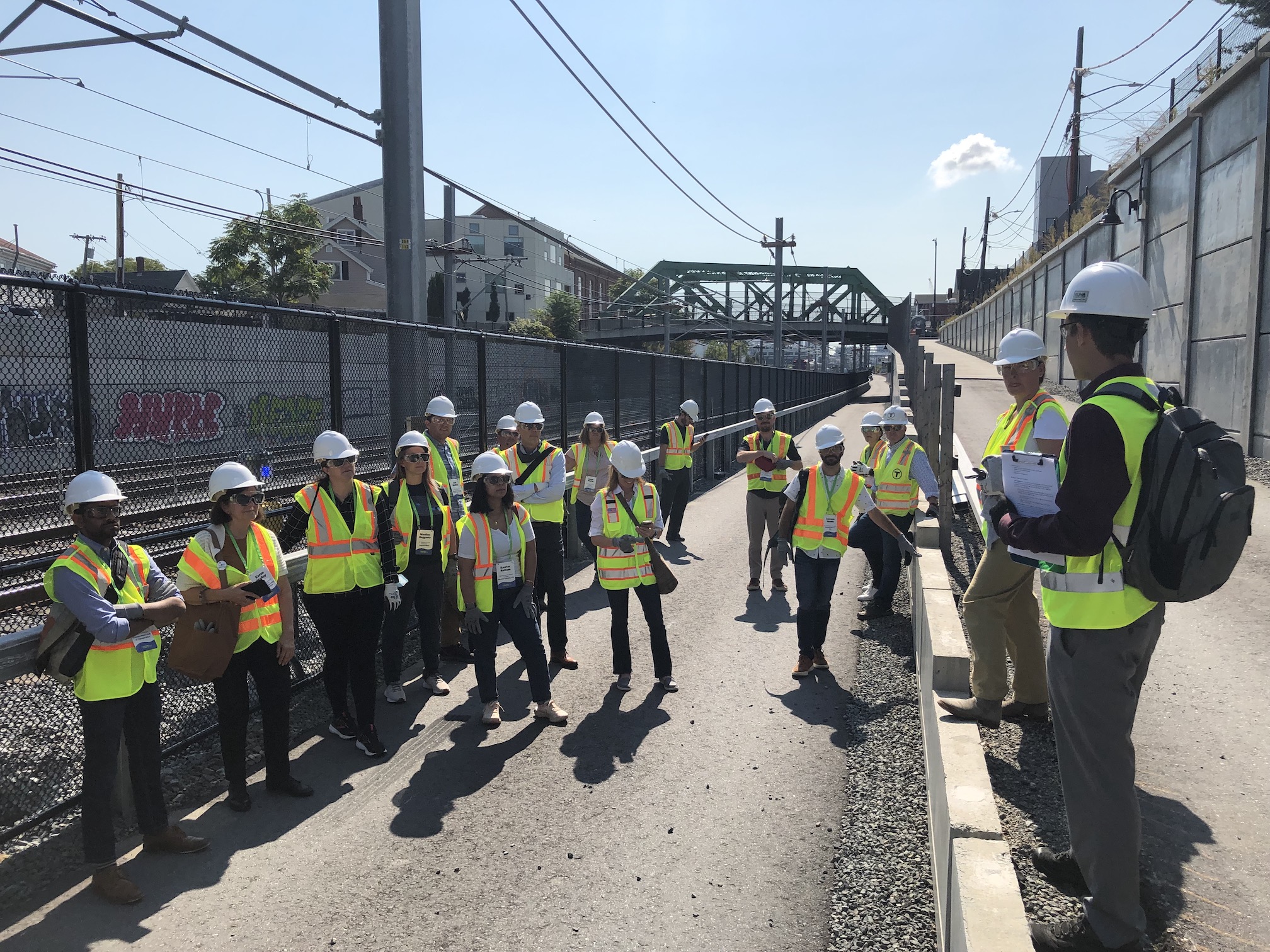 A crowd of people wearing bright construction vests and helmets listens to a man wearing a backpack at the intersection of the Community Path, which runs alongside the railroad tracks, and a second path that leads uphill to street level, above the tracks. In the distance is the Cross Street bridge.