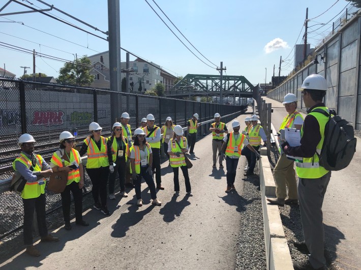 A crowd of people wearing bright construction vests and helmets listens to a man wearing a backpack at the intersection of the Community Path, which runs alongside the railroad tracks, and a second path that leads uphill to street level, above the tracks. In the distance is the Cross Street bridge.