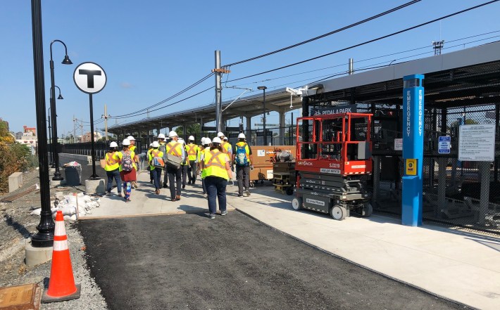 People wearing construction vests pass under a "T" sign at the construction site for the East Somerville Green Line station. To the right is a bike parking pavilion, and in the distance is the canopy of the station's train platform. A row of lights on the left edge of the photo trace the course of a ramp that leads down to Washington Street.