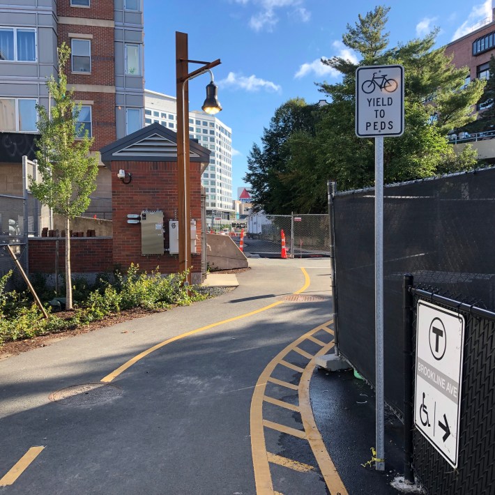 The pavement of a shared-use path meets the sidewalk of Miner Street next to a "bikes yield to pedestrians" sign. Across Miner Street, construction fencing blocks access to a new segment of trail that's under construction. In the distance is the neon Citgo sign above Kenmore Square.