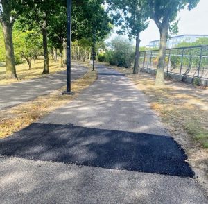 A small patch of fresh asphalt on a bike path running through the dappled shade of trees in the Southwest Corridor park