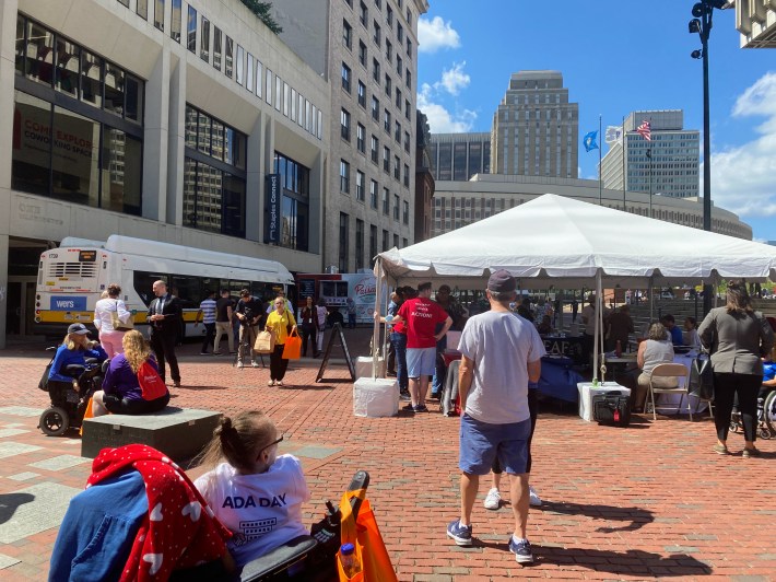 people meander through the south side of City Hall with some under a white tent and others by an MBTA bus