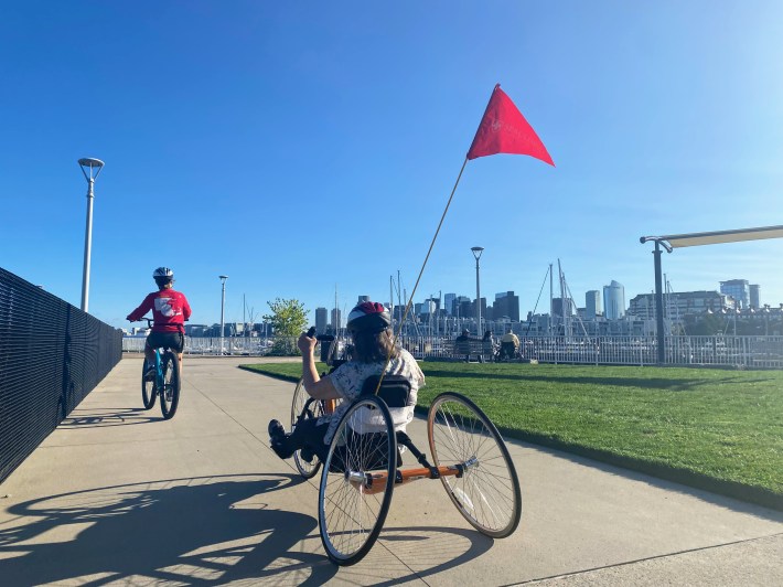 Pam Daly uses a recumbent hand cycle with a red flag pole in the back. a patch of grass is on her right and a clear blue sky ahead of her.