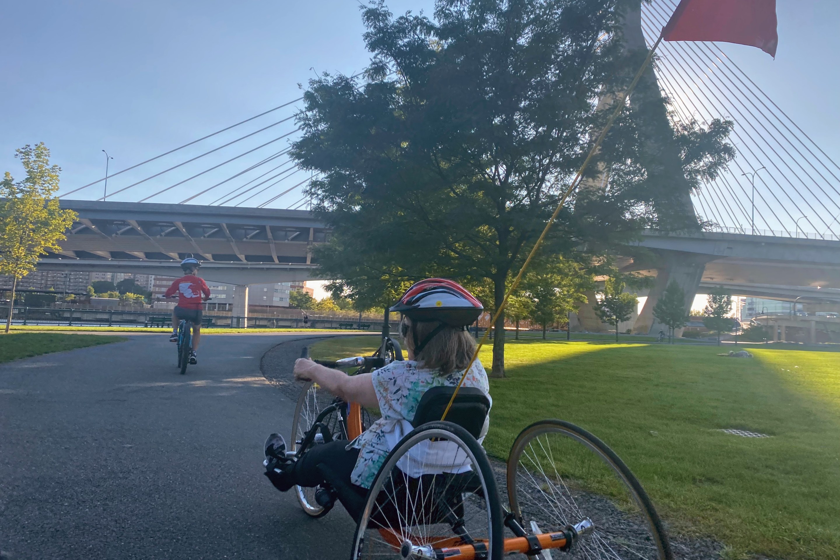 Pam Daly rides an orange recumbent bike with two wheels in the back and one in the front as she approaches the Zakim bridge with the sun behind it