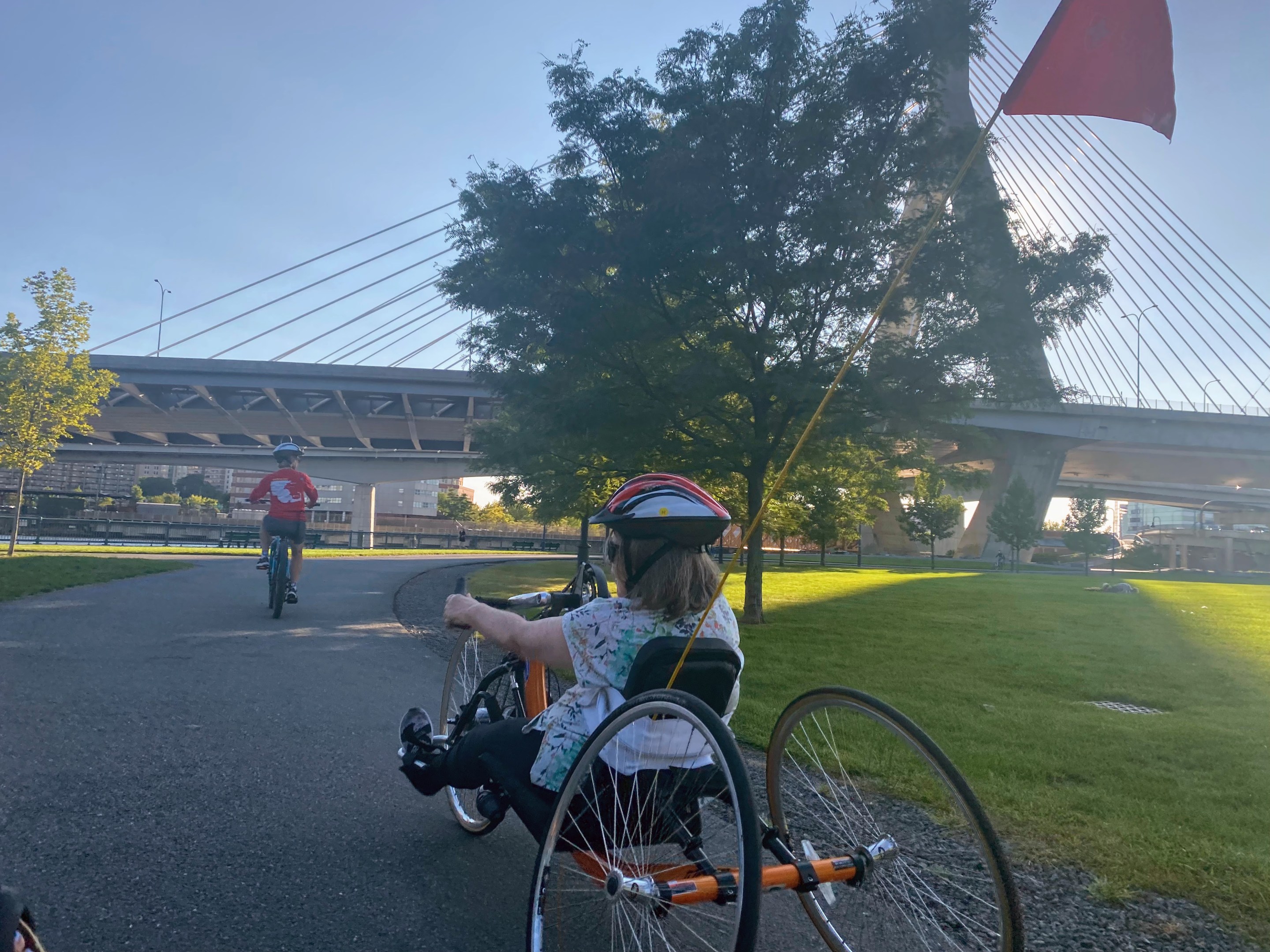 Pam Daly rides an orange recumbent bike with two wheels in the back and one in the front as she approaches the Zakim bridge with the sun behind it