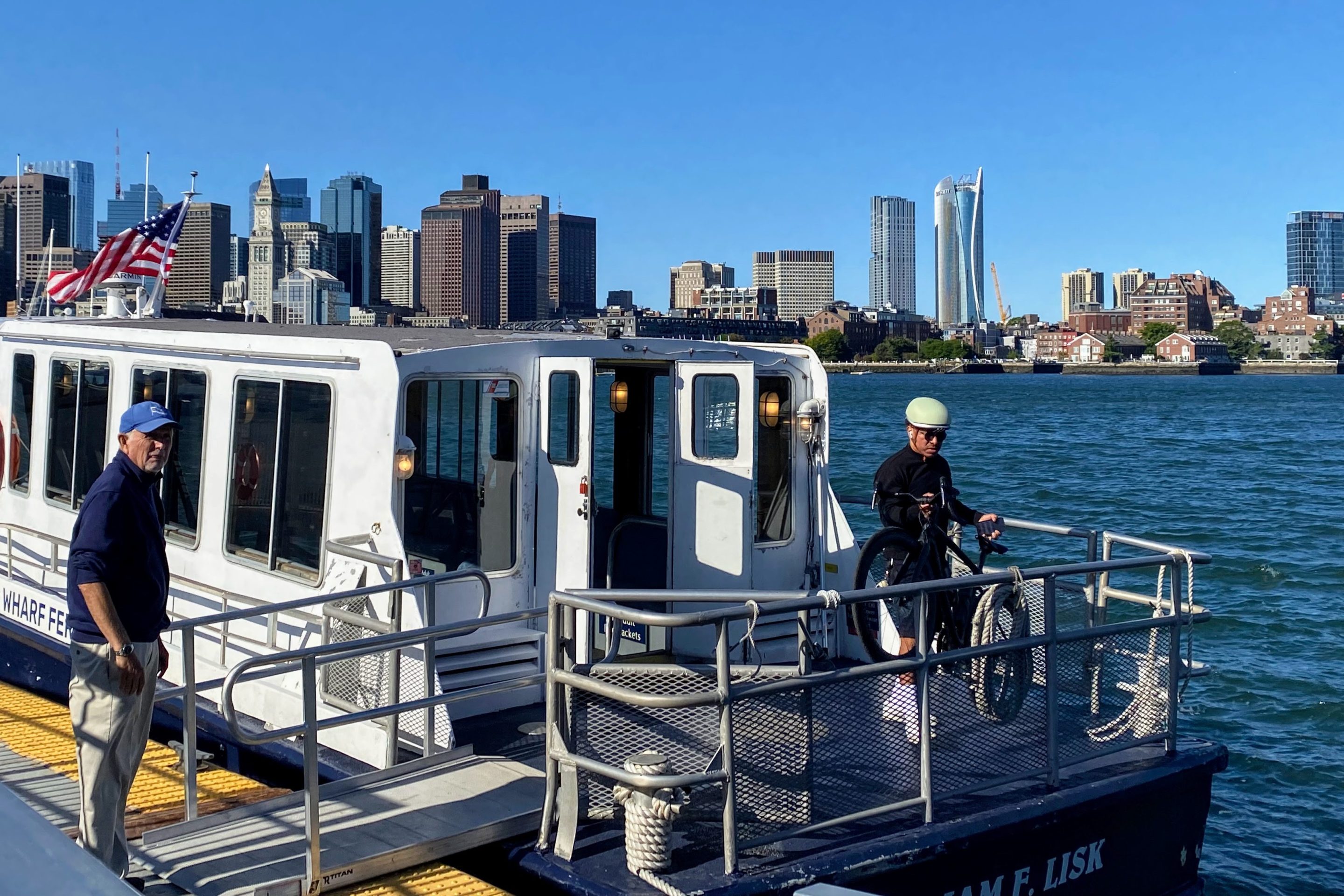A ferry attendant waits for a bicyclist to wheel his bike onboard at Lewis Wharf in East Boston. The downtown Boston skyline is visible in the background beyond Boston Harbor.