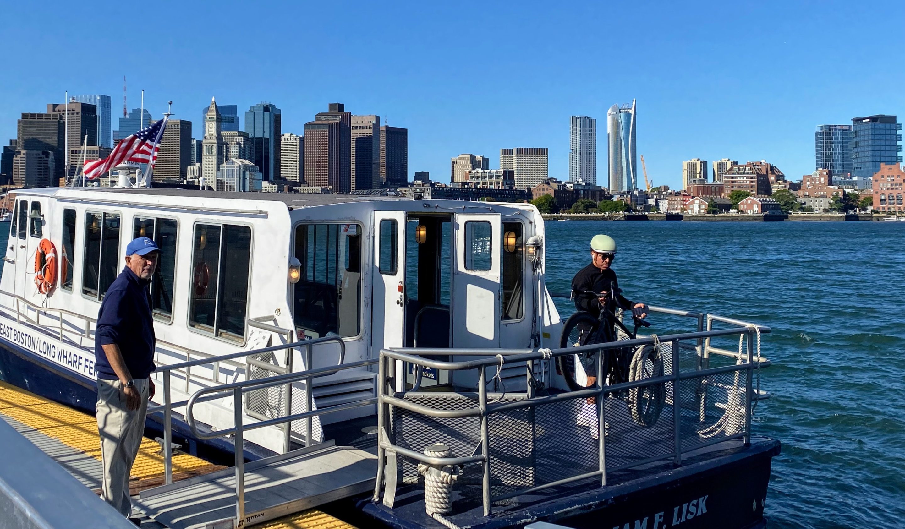 A ferry attendant waits for a bicyclist to wheel his bike onboard at Lewis Wharf in East Boston. The downtown Boston skyline is visible in the background beyond Boston Harbor.