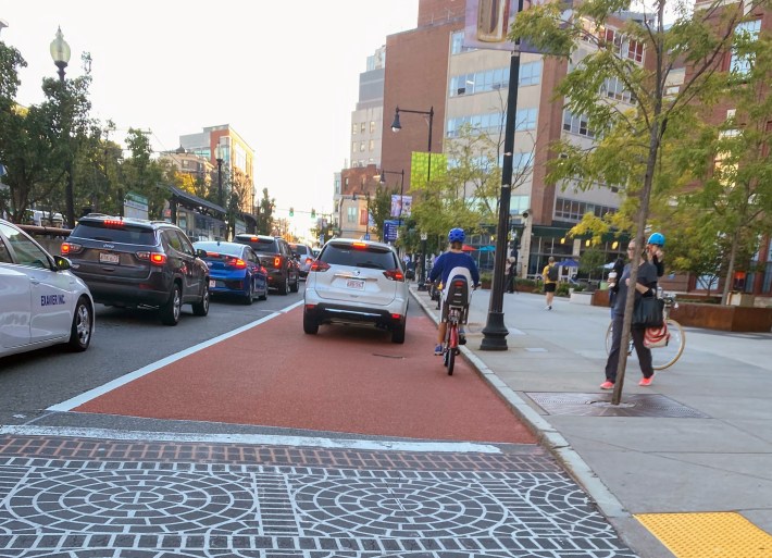 car traffic fills Huntington Avenue as pedestrians walk on the sidewalk and a bicyclists shares the bus and bike lane with a car