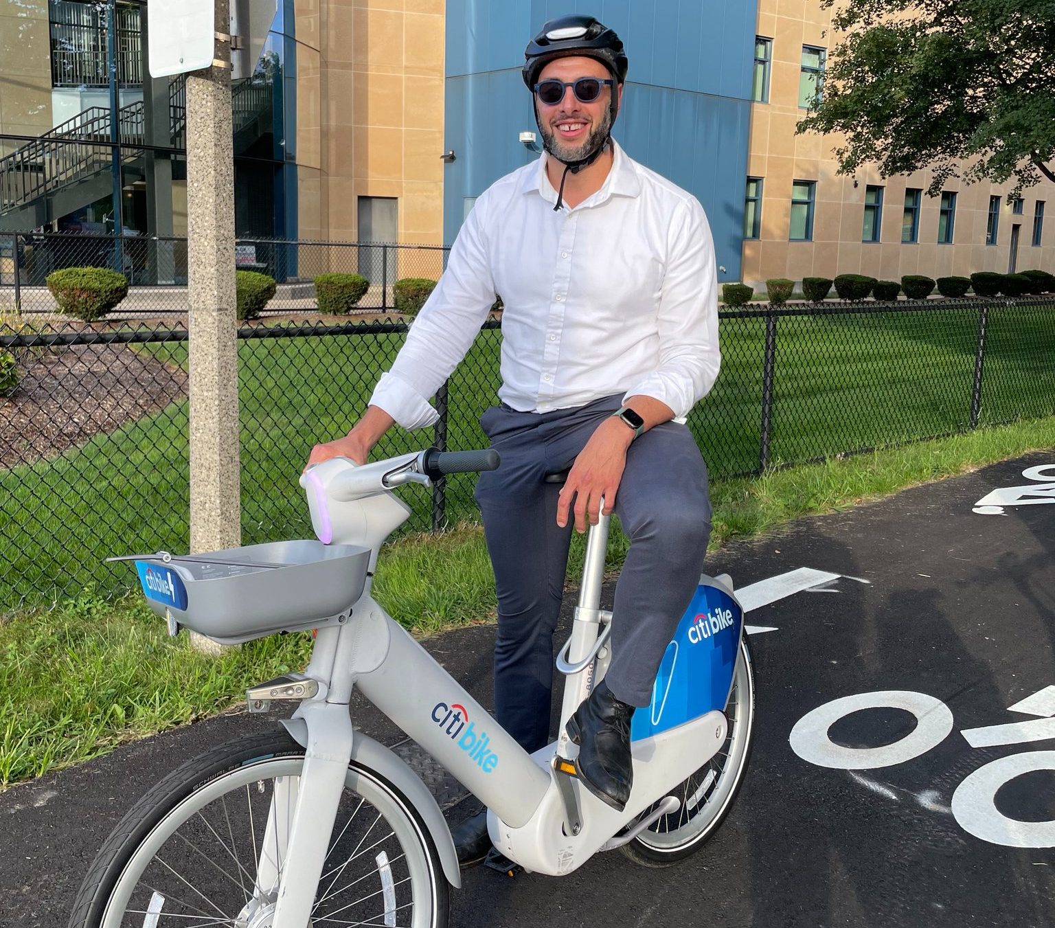 A man in a collared white shirt, sunglasses, and a helmet sits astride a sleek grey bike with the "citibike" logo on the side.